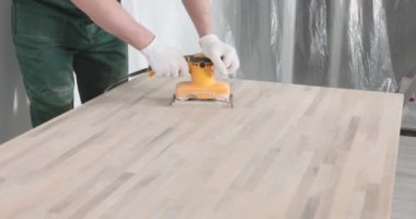 İş üniformalı bir erkek, ahşap masayı öğütme makinesiyle parlatıyor. Ağır çekimde ligneöz yüzeyleri cilalamak için profesyonel ekipman