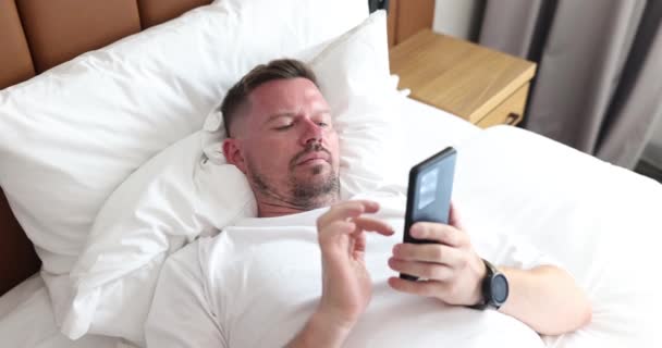 男人躺在床上 拿着智能手机读着信息 自由职业者社交网络和移动应用程序的通信 — 图库视频影像