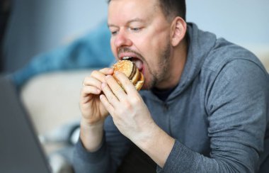 Portrait of male sitting indoors and eating big tasty hamburger full of mischievous calories and harmful cholesterol. Person consuming portion of junk food