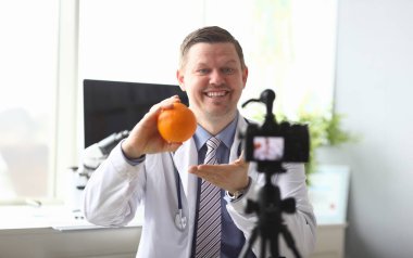 Lezzetli portakal tutan neşeli doktor portresi ve video günlüğü için yüksek teknolojili modern kameraya lezzetli organik yiyecekler gösteriyor. Beyaz üniformalı zeki bir erkek. Sağlık ve Klinik Konsepti