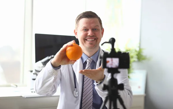 Portrait of joyful doctor practitioner holding tasty orange and showing delicious organic food to high-tech modern camera for vlog. Smart male in white uniform. Healthcare and clinic concept