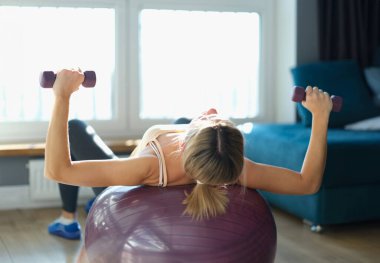 Kol halterleri ve dengeleyici topla ağırlık kaldıran genç bir kadın. Evde spor yapma konsepti