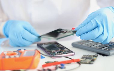 Close-up of handyman repairing broken smartphone, put cover on top, worker wear gloves. Workplace full with equipment. Repair service, fix, handy concept