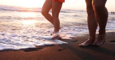 Siyah kumlu sahilde yürüyen çift. Köpüklü deniz dalgaları turistlerin bacaklarını yıkıyor. Gün batımında aydınlanmış olarak denizin önünden geçiyorlar.