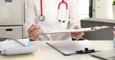 Steteskoplu bir doktor hastaya iş yerinde otururken imzalaması için kalem ve pano veriyor. Tedavi belgesinin imzalanması yavaş çekim