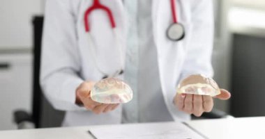 Steteskoplu doktor, iş yerindeki masada oturan göğüs implantları için seçenekler gösteriyor. Klinikte yavaş çekimde estetik ameliyat ve yeniden yapılanma modelleri kavramı