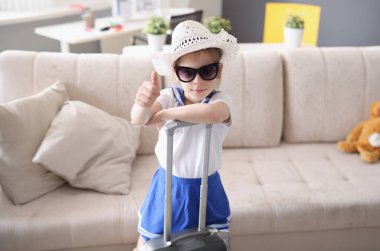 Şapkalı ve güneş gözlüklü küçük kız bavuluyla ayakta duruyor ve baş parmağını kaldırıyor. Çocuklar yaz tatili ve tatil tavsiyeleri konsepti