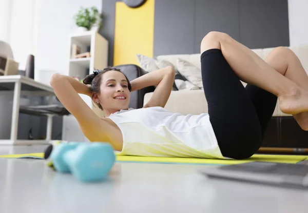 Young woman toned abs on rug at home while watching video guide on laptop. Home workouts and online fitness classes concept