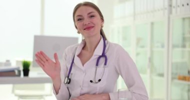 Kameraya bakıp seyircilere el sallayan gülümseyen kadın doktorun portresi. Hasta ile sanal çevrimiçi görüşmede uzaktan danışmanlık yapan genç bir kadın doktor.