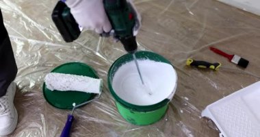 İşçi kapta beyaz sıvı boyayı karıştırıyor ve yerde folyoyla kaplanmış endüstriyel karıştırıcı var. Ressam dairede bakım için gerekli malzemeleri hazırlar.