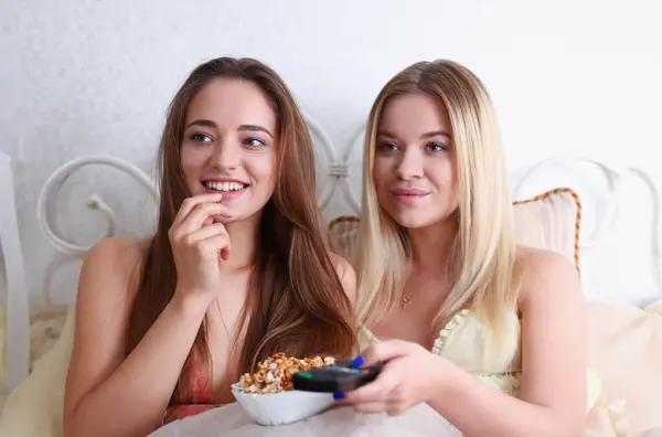 Two happy smiling girlfriends eat popcorn in bed watching TV show early morning holding remote control in hand portrait. Joy cozy tenderness lifestyle football match happiness or tv series concept