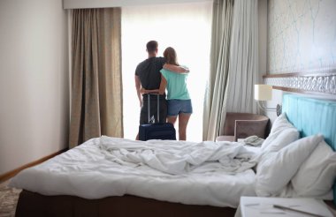 Erkek ve kadın, bavulun yanında pencerenin yanında birbirlerine sarılıyorlar. Romantik gezi konsepti nereye gider?