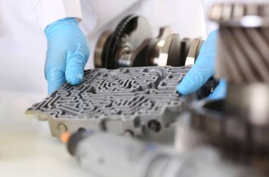 Otomobil tamircisi otomatik vites kutuları elinde mavi koruyucu eldivenlerle duruyor. Hidroblok detayı tanıları kurutur ve detaylı test iletimini yakınlaştırır.