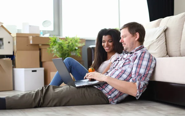 Portrait of couple sitting on fluffy carpet in new apartment. Woman and man entertaining with modern laptop. Empty boxes in room. Moving day and real estate concept