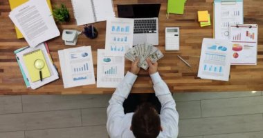 İş yerindeki bir masada çalışan bir işadamı el ile para sayıyor ve gelir tablolarını ve grafiklerini analiz ediyor. İş stratejisi ve finansal gelir ve zenginlik duygusu
