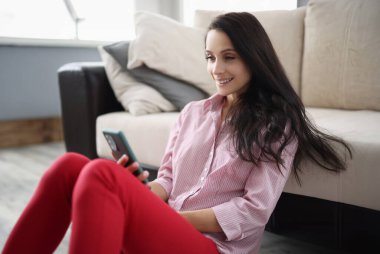 Happy pleasant woman resting at home holding smartphone. Smiling young lady chatting on social networks watching funny videos and using mobile apps at home