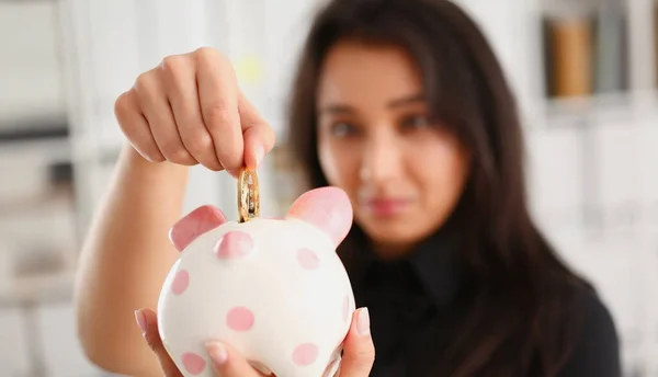 A young smiling chinese woman holding bitcoin coin in his hand thrusts it into piggy bank form of pink piggy. Saving storage theme in crypto currency.