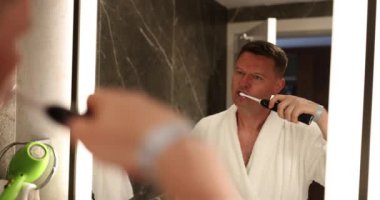 4K filminde beyaz bornozlu bir adam elektrikli diş fırçasıyla dişlerini fırçalıyor. Ağız temizliği kavramı