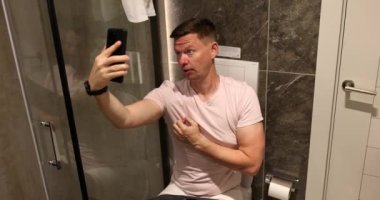 Adam tuvalette oturup cep telefonu kamerası 4K filminde selfie çekiyor. Ev kavramından sıkıldığınızda