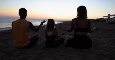 Aile siluetleri gün batımında yoga yapıyor. Kumsalda huzur ve rahatlama.