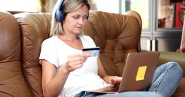 Kulaklıklı bir kadın dizüstü bilgisayarla koltukta oturuyor ve kredi kartıyla online alışveriş yapıyor. Çevrimiçi mağazada çevrimiçi ödeme