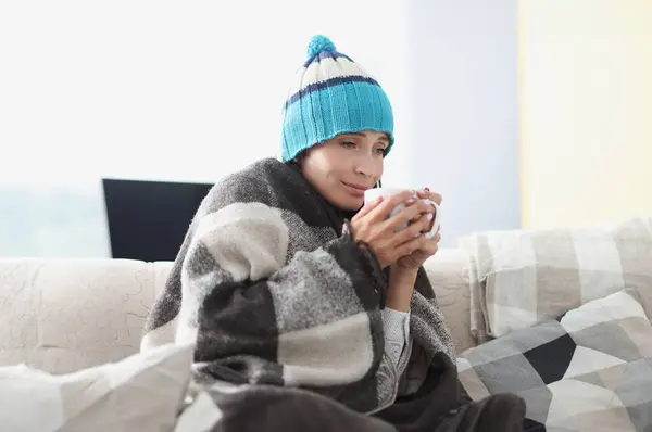 Chilled woman on couch in blanket and hat holds hot cup. Drinking for colds concept