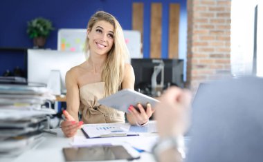 Ofisteki çalışma masasındaki gülümseyen iş kadını tablet tutuyor ve iş göstergelerini inceliyor. Kârlı işbirliği ve etkileşim konsepti