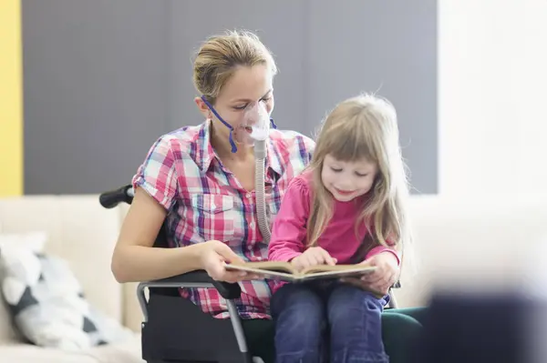 Woman with an oxygen mask and in wheelchair is reading book with little girl. Communication with loved ones in case of serious illnesses concept