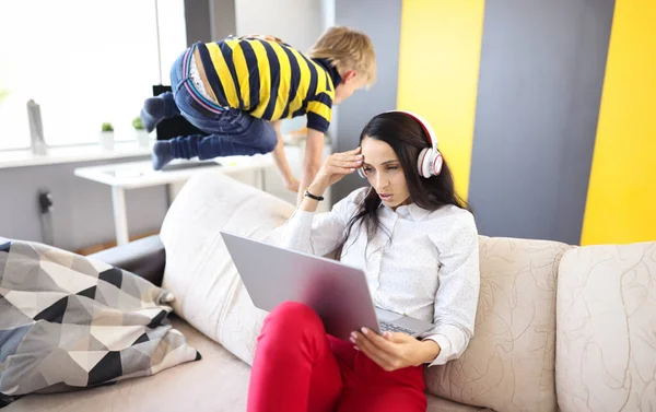 Woman in headphones and laptop sits on sofa next to child pampering. How to combine remote work at home and children concept