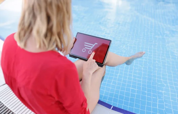 Top view of woman using tablet for online shopping, red background with shopping cart. Shop from any place, woman on holiday in pool. Shopaholic concept