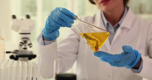 化学科学者は透明なボトルから実験室のフラスコに水を注ぐ 化学実験室の閉鎖の透明性のための黄色い液体かオイルが付いているフラスコを見る科学者の化学者 — ストック動画