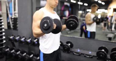 Formda ve kaslı adam spor salonunda ağır dambıllarla antrenman yapıyor. Erkek vücut geliştirici beden eğitimi alıyor ve dambılı elle kaldırıyor.