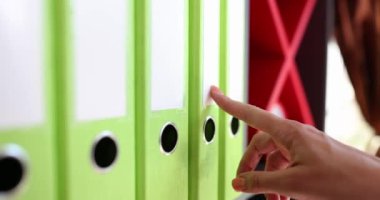 Kadın elleri raftaki klasörlere dokunmuş, yakından, yavaş çekimde. Mali belge yönetimi, arşiv