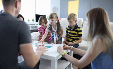 Yetişkinler ve çocuklar oyun kartlarının bulunduğu masanın etrafında oturuyorlar. Kız eliyle kart için uzanıyor. Ev kavramındaki çocuklar ve yetişkinler için aile oyunları