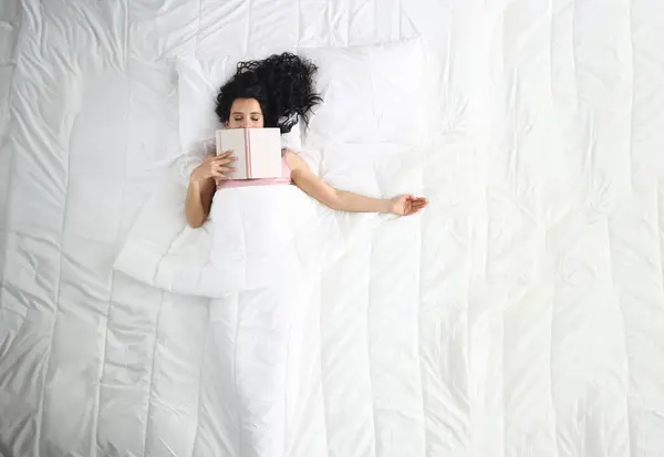 Woman lies on white bed with closed eyes book lies on her face. Relax and tranquility before bed concept
