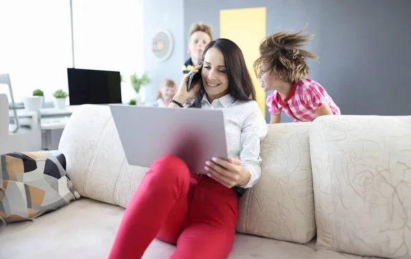 Woman is sitting on couch holding laptop and talking on smartphone next to the children are playing. Remote work in coronavirus in family concept