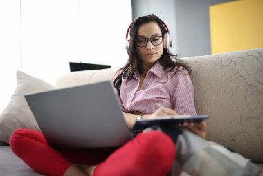 Elinde dosya olan kulaklıklı bir kadın dizüstü bilgisayarla kanepede oturuyor. Çalışanların uzaktan çalışma konseptine geçişi