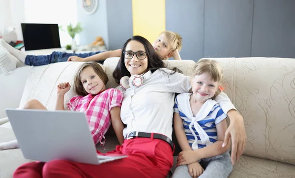 Woman is sitting on sofa with two girls and a boy is standing next to laptop. Teaching children online in coronavirus concept