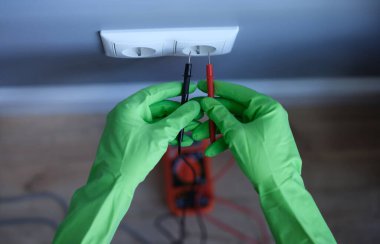 Lastik eldivenli bir zanaatkar, yakın çekimde elektrik prizinin yanında test cihazı tutuyor. Elektrik kavramı ile çalışırken güvenlik