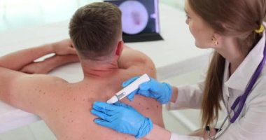 Elektronik cihaz erkek hastanın sırtında papilloma gösteriyor. Müşteri, kadın doktor tarafından ağır çekimde incelenen hasarlı sırt derisini gösteren monitöre bakıyor.