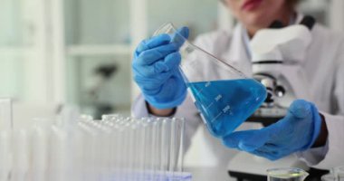 Odaklanmış kadın bilim adamı, laboratuardaki iş yerinde mikroskop yakınında mavi sıvıyla matarayı inceliyor. Araştırma için steril dayanak yavaş çekim