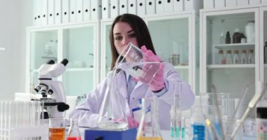 İş üniformalı kadın bilim adamı cam kaptan şeffaf sıvıyı masada mikroskop ile oturarak şişeye boşaltıyor. Uzman yavaş çekim tarafından keşfedilme