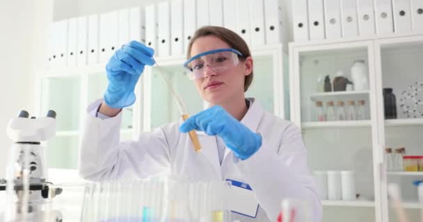 ワークユニフォームの女性化学者は 顕微鏡でテーブルでサンプルを調べるスティック付きのガラスチューブの液体を攪拌します 実験室でのプロの仕事 — ストック動画