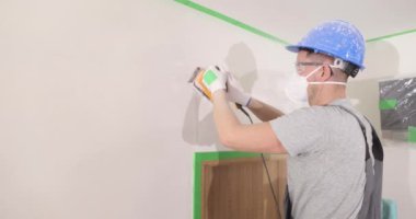 Builder in helmet and goggles polishes plaster spot on wall in restored room. Employee fixes holes and trenches on drywall panel with tool
