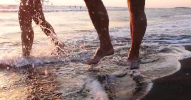 Deniz dalgaları tarafından yürüyen çift bacakları karanlık kumlu sahilde. Köpüklü deniz dalgaları turistlerin bacaklarını yıkıyor. Gün batımında aydınlanmış olarak denizin önünden geçiyorlar.