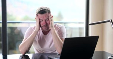 Evde dizüstü bilgisayarın önünde yorgun bir adam kafasına yavaşça masaj yapıyor. Uzak çalışma, stresli durum