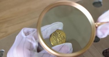 Hands in gloves hold an old coin and a magnifying glass, close-up. Estimated value from an antique dealer