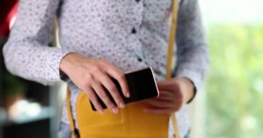 A girl in a shirt puts a smartphone in a leather purse, close-up. Woman with a bag and phone in the office