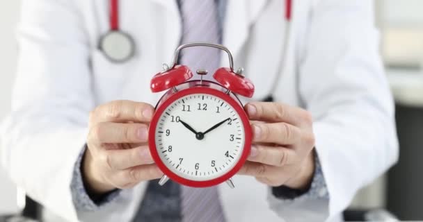 若い笑顔の医者は朝１０時に赤い目覚まし時計を持っている 勤務日医療従事者の概念 — ストック動画