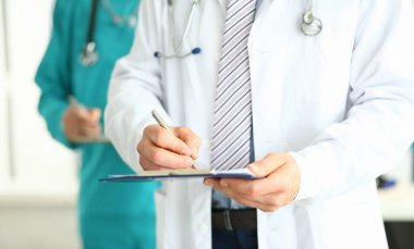 Not defterine bir şeyler yazan profesyonel hekimlere odaklanın. Klinik ofisinde duran akıllı doktor. Laboratuvar önlüğü ve klas kravat giyen bir adam. Tıbbi tedavi ve sağlık kavramı